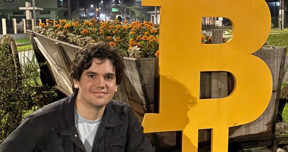 Neto Lócio, co-founder of ChampQuest and SEO strategist, posing beside a large Bitcoin symbol in an urban outdoor setting.